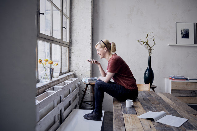 Stockfoto, lizenzfrei, Frau sitzt auf Schreibtisch im Loft und benutzt Mobiltelefon