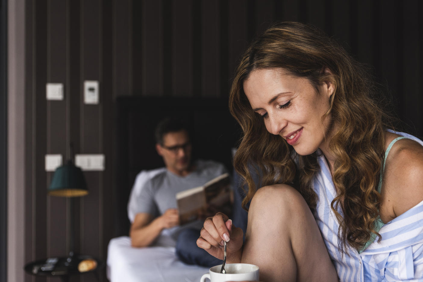 Smiling woman with cup of coffee in bedroom with man in background
Royalty free image