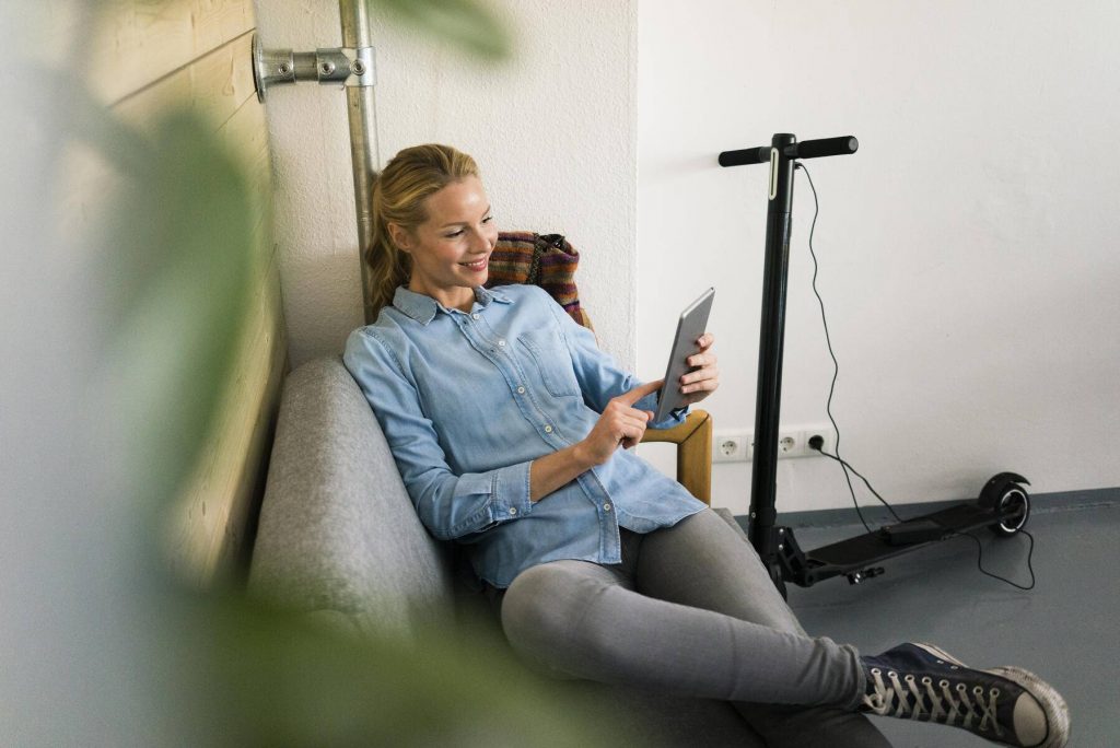 Young woman sitting on couch in a coffe shop, using digital tablet, e-scooter charging in background - JOSF03339