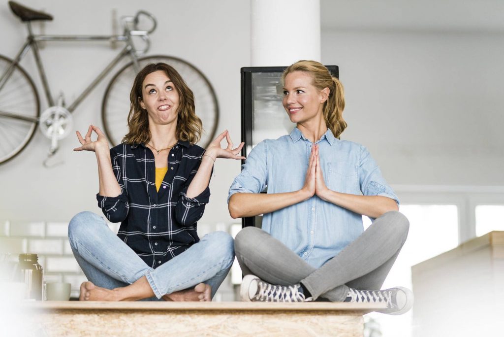 Two young women sitting cross-legged on the counter of their coffee shop, one meditating , other pulling funny faces - JOSF03435