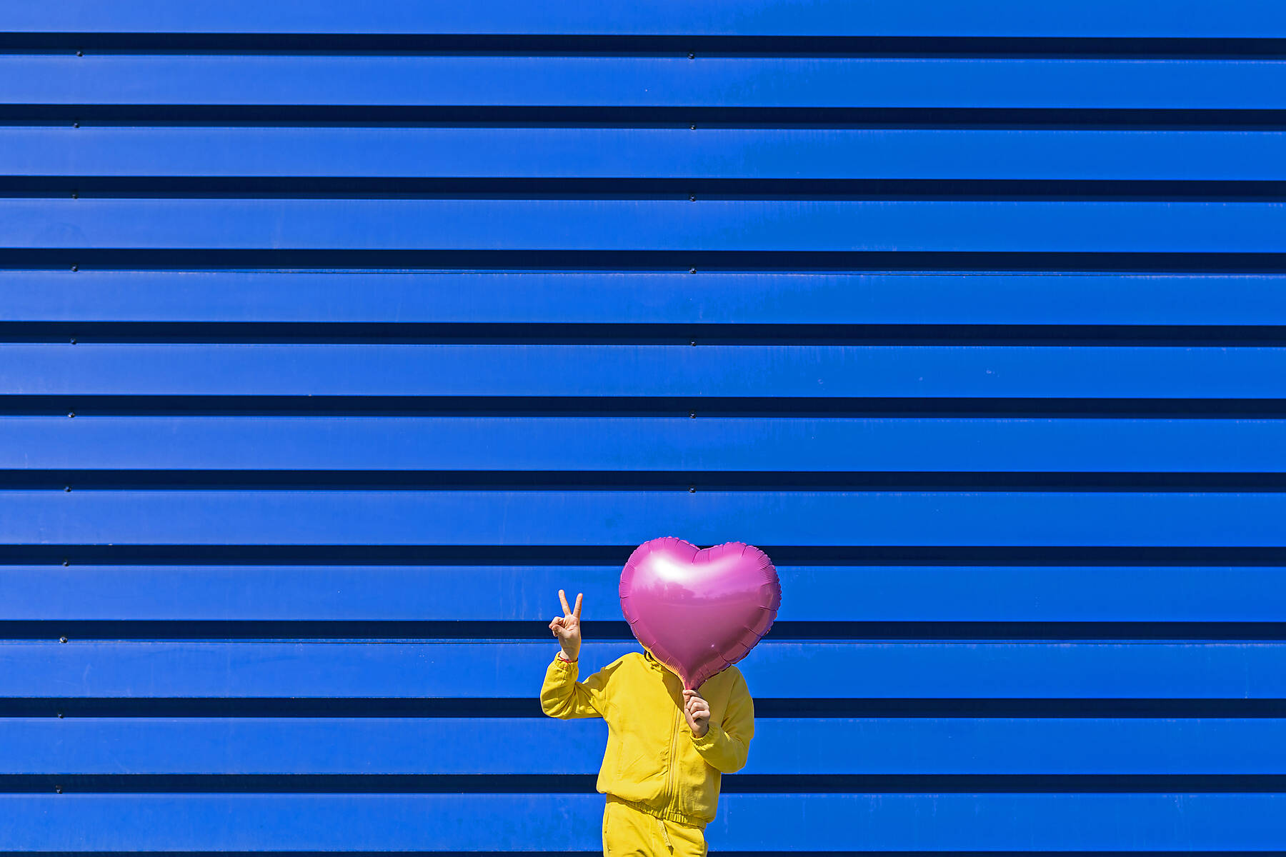 royalty-free, stock image, Little girl standing in front of blue background hiding behind pink balloon and showing victory sign