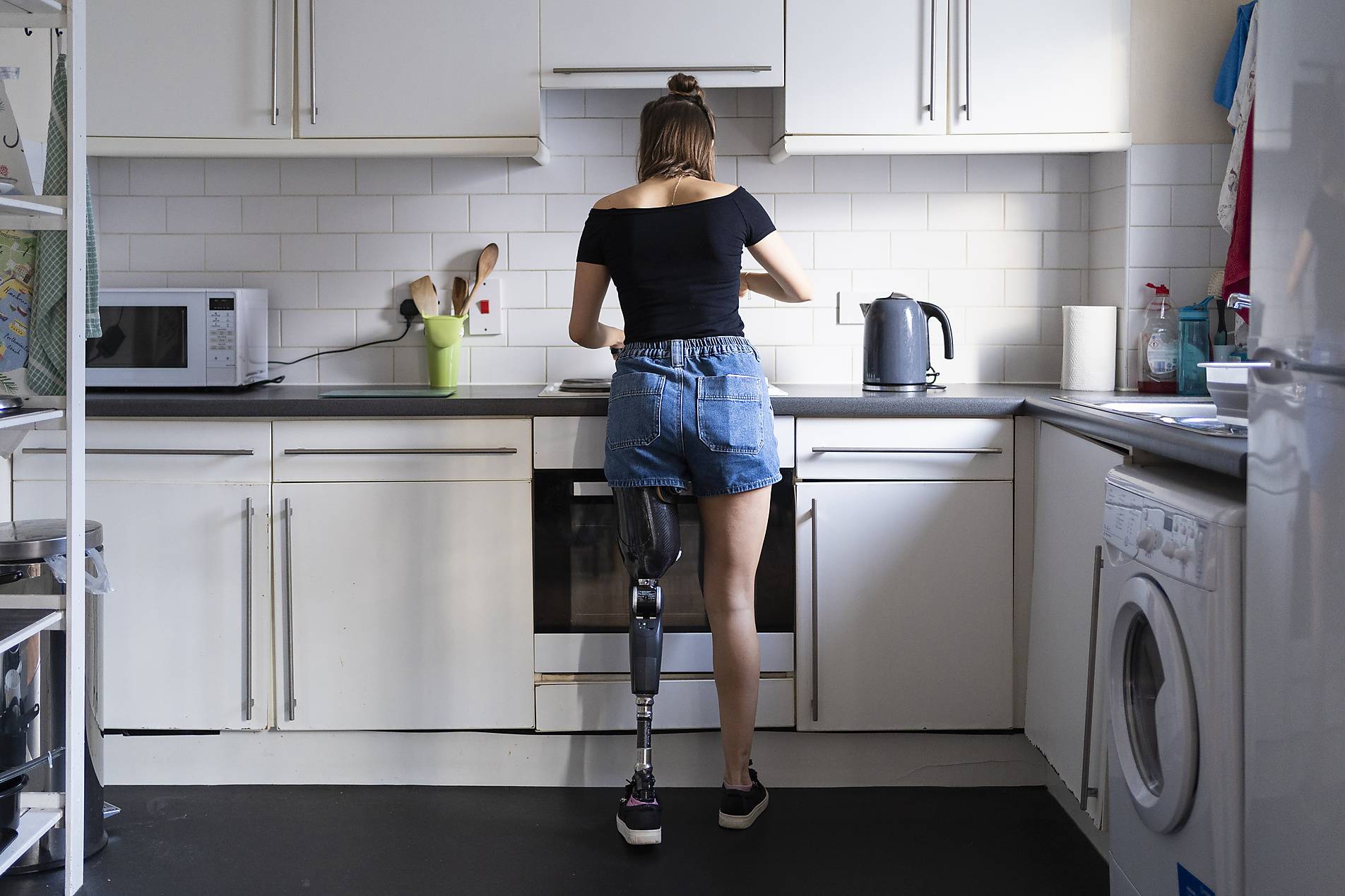 Stock photo, royalty-free, Rear view of young woman with leg prosthesis in kitchen at home, Diversity