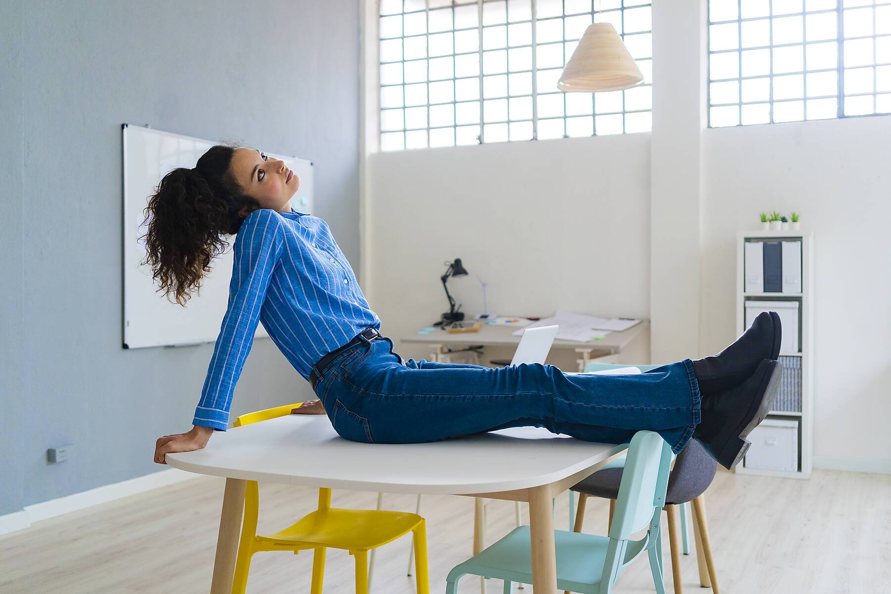 Young businesswoman with legs crossed at ankle contemplating while sitting on desk in office