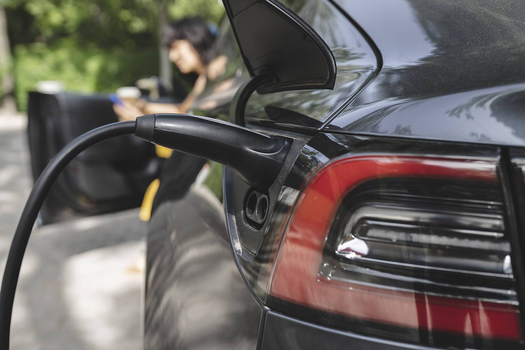 Stockfoto, lizenzfrei, Kabel im Stecker eines Elektroautos an einer Ladestation mit einer Frau im Hintergrund