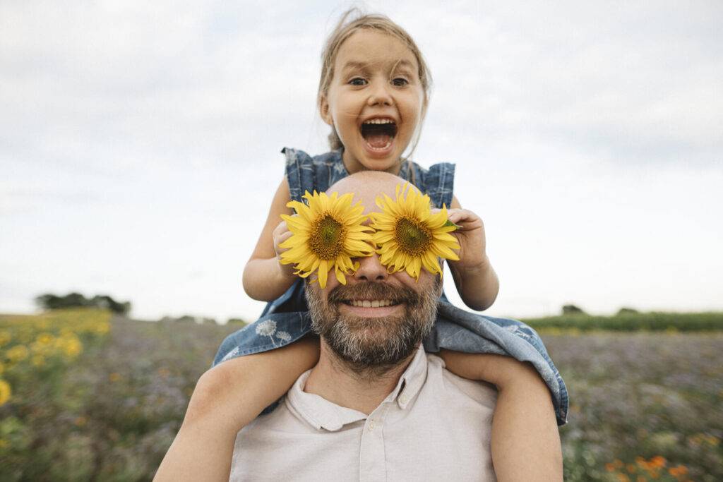 stockfoto, lizenzfrei, Sonnenblumen bedecken die Augen eines verspielten Mannes mit Tochter auf einem Feld