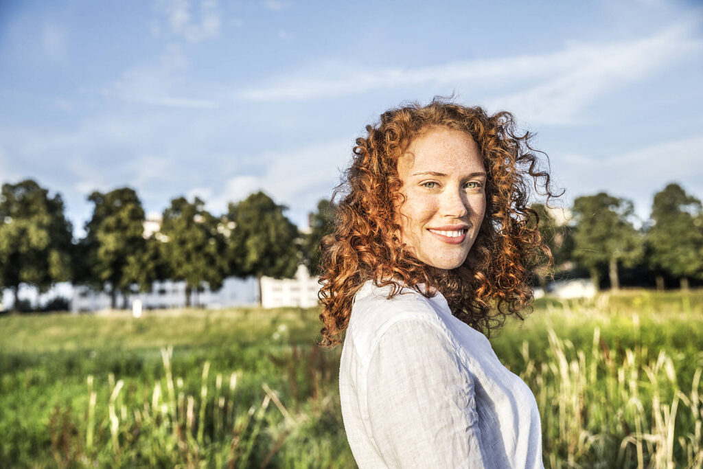 Stockfoto, lizenzfrei, Deutschland, Köln, Porträt einer lächelnden jungen Frau mit lockigem roten Haar in der Natur