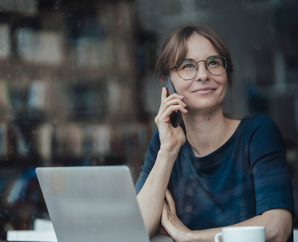 Stockfoto, lizenzfrei, Lächelnde weibliche Fachkraft, die wegschaut und mit einem Smartphone in einem Café spricht, gesehen durch ein Glasfenster