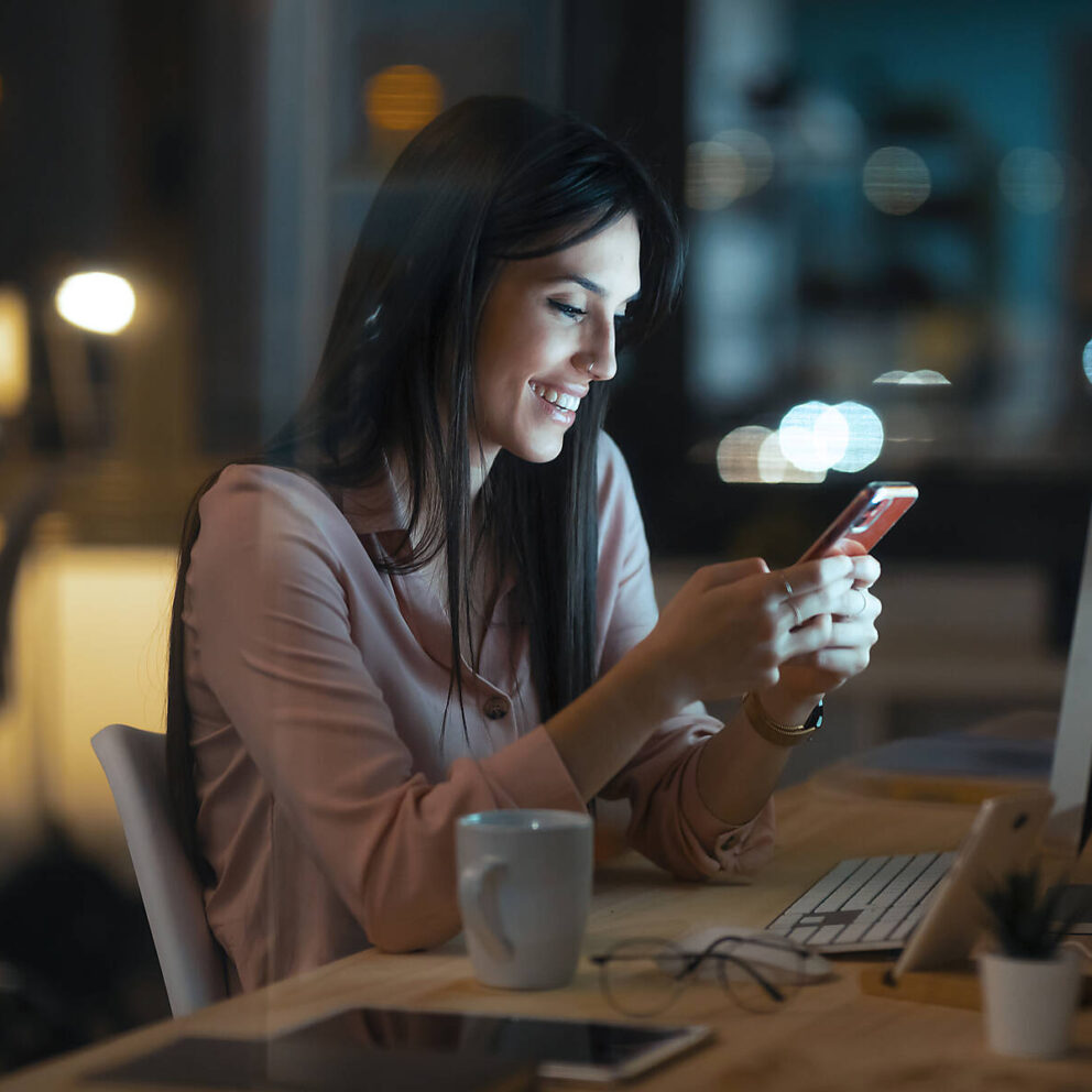 Stockfoto, lizenzfrei, Lächelnde junge Frau sitzt im Büro am Schreibtisch und schaut auf ein Smartphone