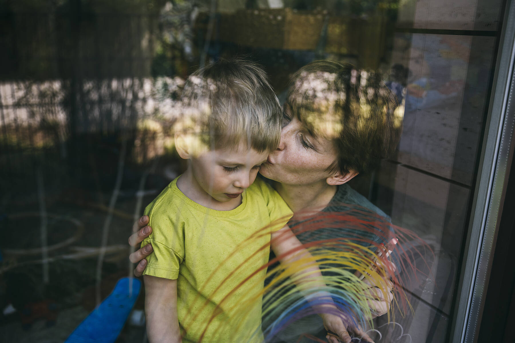 Mutter küsst ihren kleinen Sohn hinter dem Fenster mit gezogenem Regenbogen