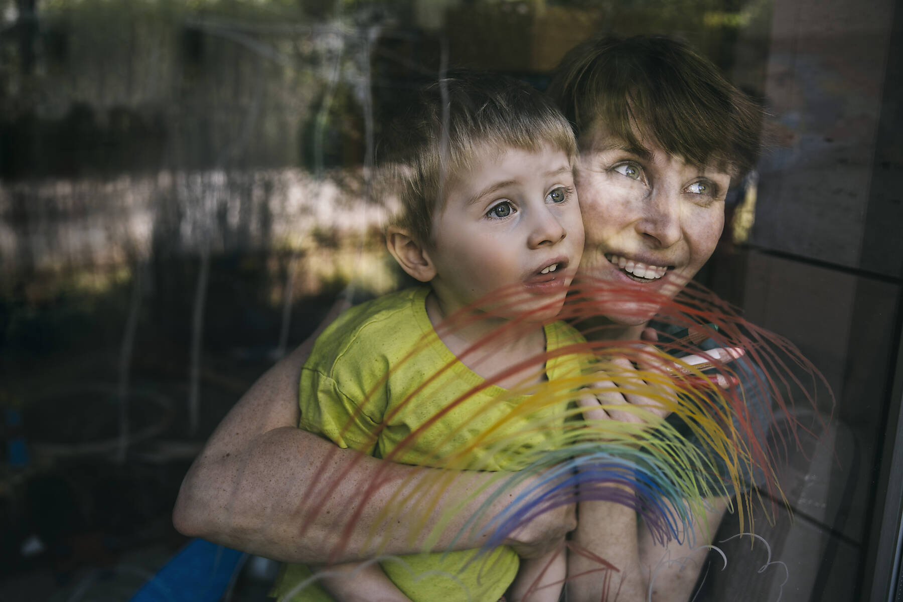 Porträt einer glücklichen Mutter und ihres kleinen Sohnes, die mit gezeichneten Regenbogen aus dem Fenster schauen