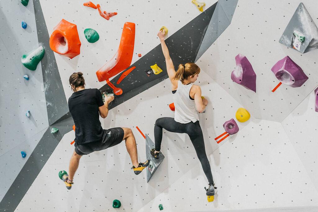 Stockfoto, lizenzfrei, Rückenansicht eines sportlichen Paares in Sportkleidung, das beim gemeinsamen Training in einem hellen, modernen Fitnessstudio eine Kunstwand mit bunten Griffen erklimmt