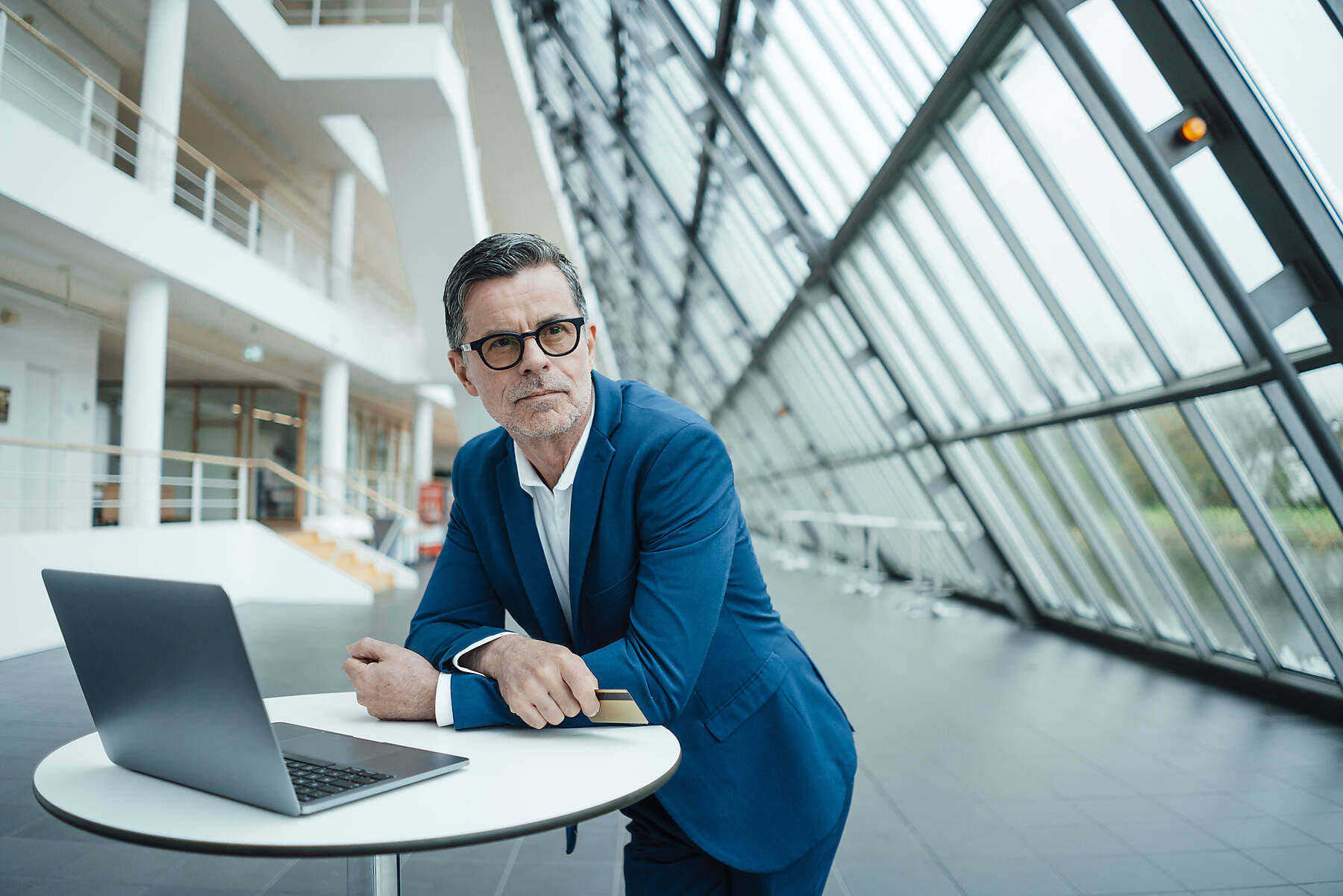 Stockfoto, lizenzfrei, Nachdenklicher Geschäftsmann mit Kreditkarte, der sich an einen Laptop auf einem Tisch im Büro lehnt