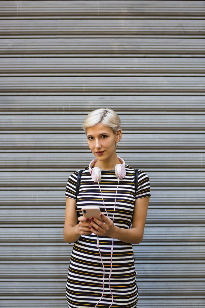 Stockfoto, lizenzfrei, Porträt einer jungen Frau mit Kopfhörern und Smartphone im gestreiften Kleid vor einem Rolltor