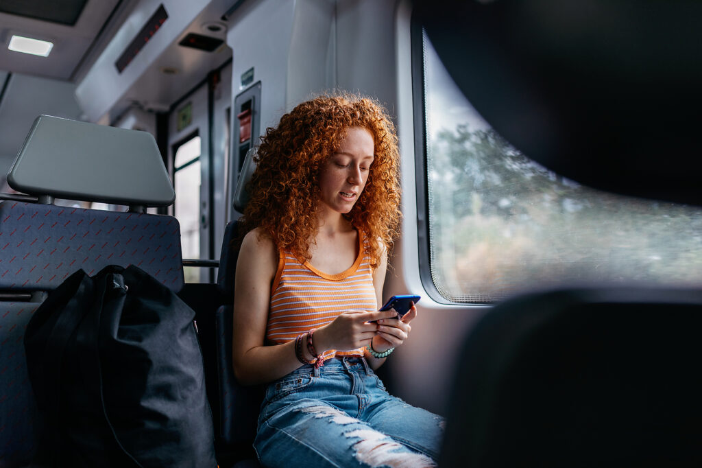 Stockfoto, lizenzfrei, Interessierte Frau mit lockigem Haar in zerrissenen Jeans, die während einer Zugfahrt tagsüber eine Textnachricht auf ihrem Mobiltelefon sendet