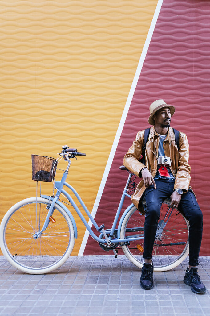 Stockfoto, lizenzfrei,vertikale Bilder, Stories, Storytelling, mobile-first, Ganzkörperaufnahme eines ruhigen Afroamerikaners mit Hut, der in die Ferne schaut, während er auf einem Fahrrad auf dem Bürgersteig in der Nähe einer bunten Wand sitzt