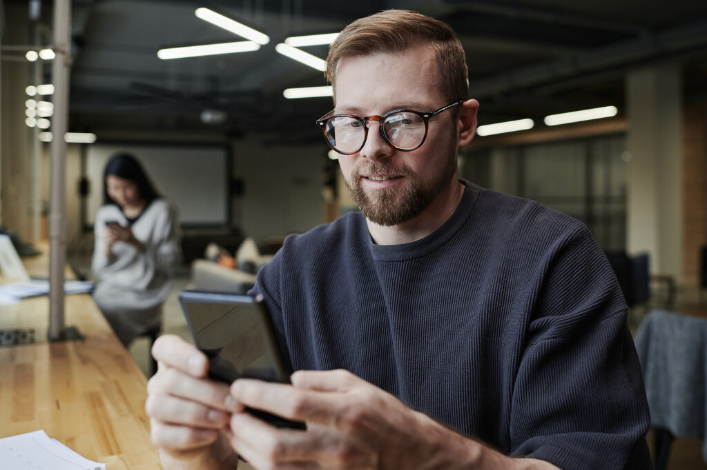 Stockfoto, lizenzfrei, Lächelnder Geschäftsmann mit Brille, der ein Smartphone im Büro benutzt