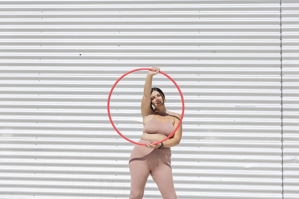 stockfoto, lizenzfrei, Kurvige Frau hält Plastikreifen vor einer Wellblechwand, body positivity,