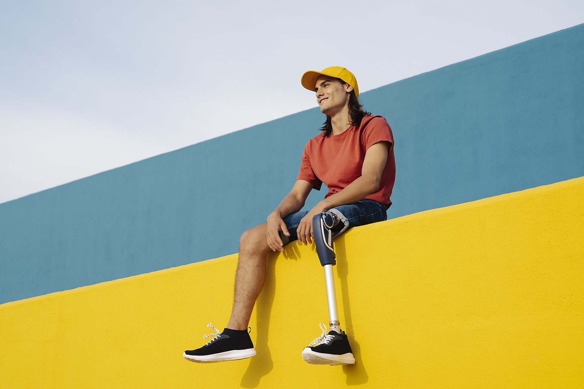Junger Mann mit Beinprothese lächelt, während er an der Wand vor klarem Himmel sitzt, body positivity,