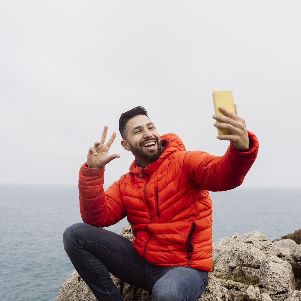 stock image, royalty-free, jose carlos ichiro taking a selfie in front of the sea, gesturing the peace sign, orannge jacket