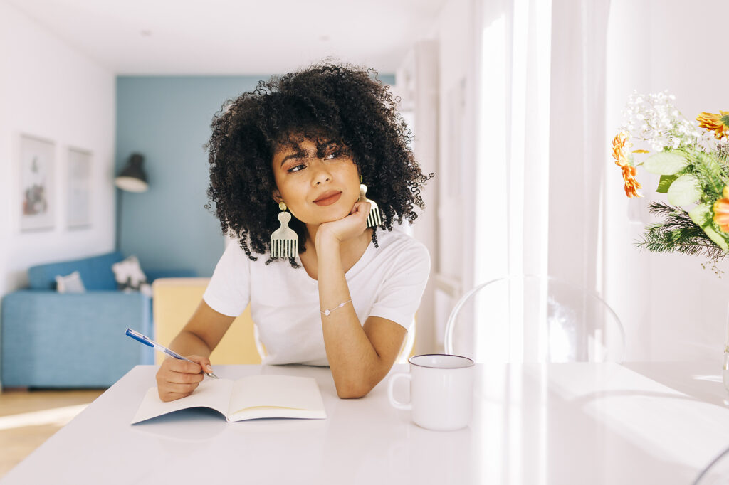 Stockfoto, lizenzfrei, Porträt einer jungen Frau mit lockigem Haar, die zu Hause mit einem Notizbuch an einem Tisch sitzt, Alexandra C Ribeiro