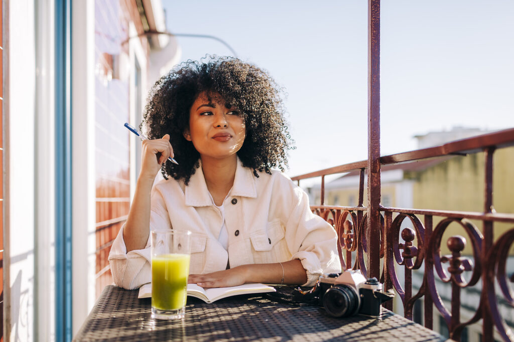 Stock image, royaly-free, Alexandra C Ribeiro, Portugal, Smiling young woman with curly hair sitting at table on balcony with notebook, smoothie and camera