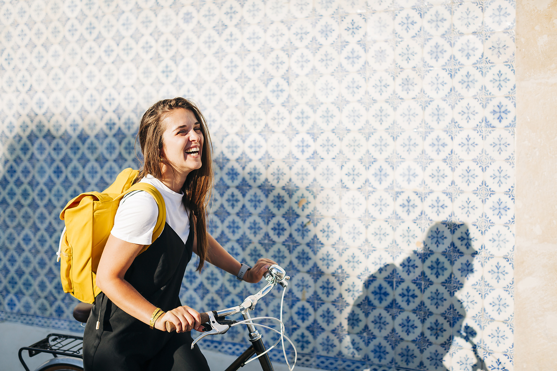 Stock image, royalty-free, Cheerful woman with yellow backpack riding bicycle by tiled wall in city on sunny day