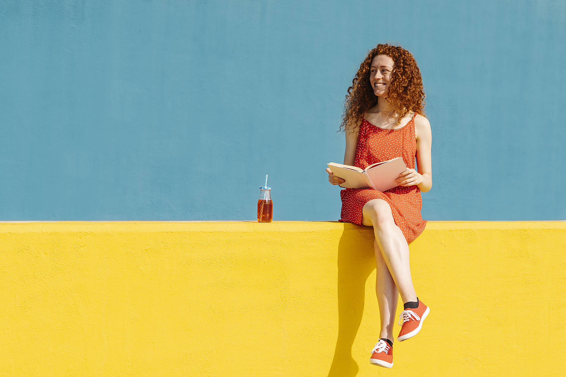 Stock image, royalty-free, Joan Corominas, Smiling woman sitting with book while looking away on retaining wall