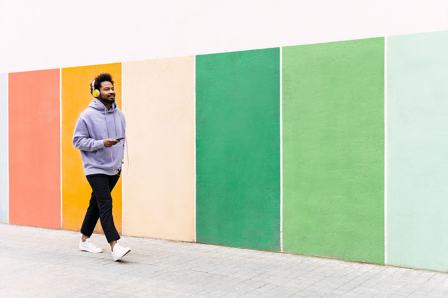 stock image, royalty-free, Man holding mobile phone listening music through headphones walking on footpath by colorful wall