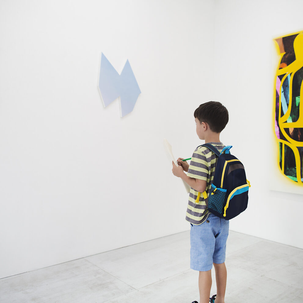 Stock image, royalty-free, westend61, future, eduction, school, Boy with short black hair wearing backpack standing in art gallery, holding pen and paper, looking at modern painting.