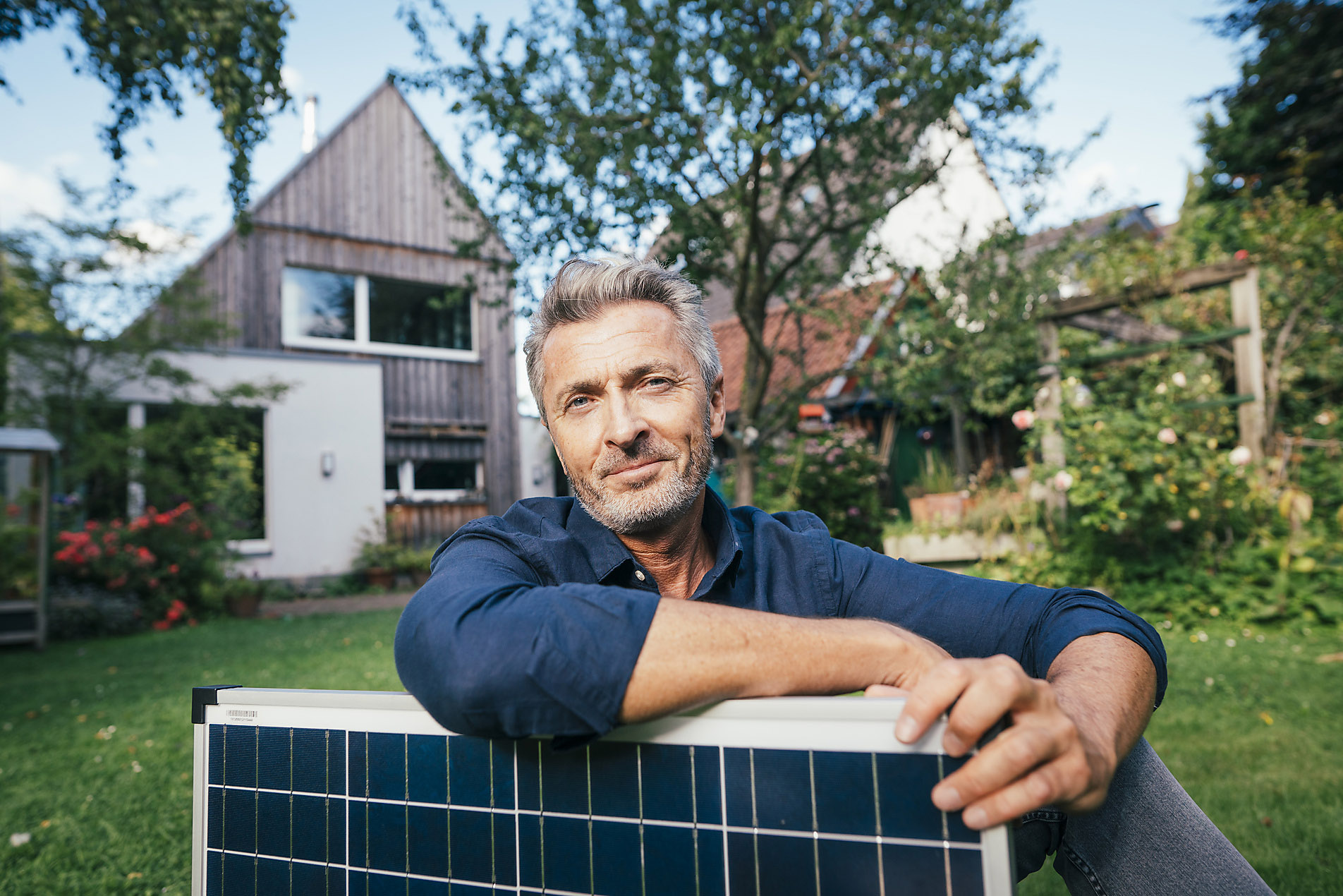 Smiling mature man sitting with solar panel in front of house, Stock photo, royalty free, sustainability, resilience, solar energy, trend report 2023, visual trends