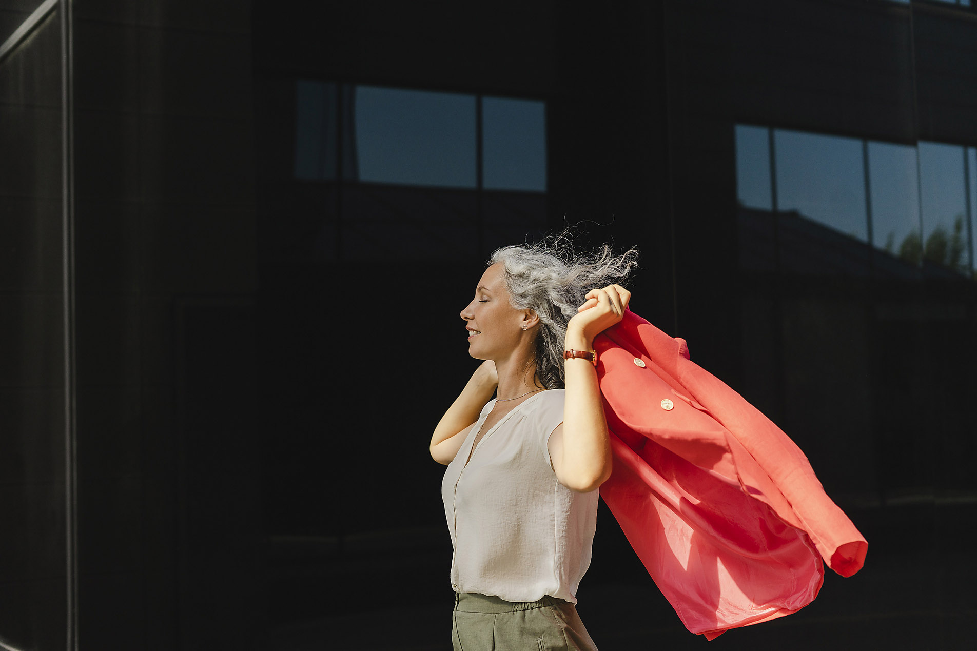 Smiling business woman with closed eyes holding a red blazer in front of a wall on a sunny day, stock photo, royalty free, resilience, visual trends, trend report 2023
