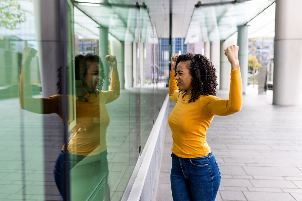 Woman looking at herself flexing muscles looking reflection on glass wall, inner strenght, resilience, westend61