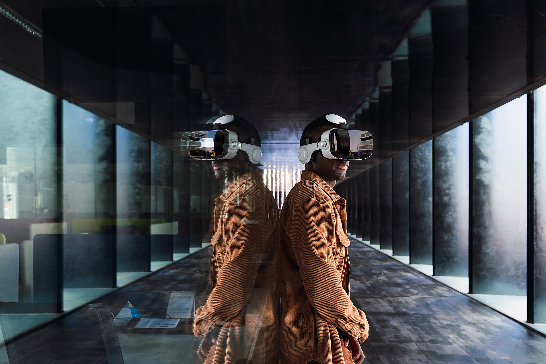 Side view of African American male in modern VR goggles standing backwards a glass wall with reflection while exploring virtual reality, metaverse, stock image, royalty-free, westend61