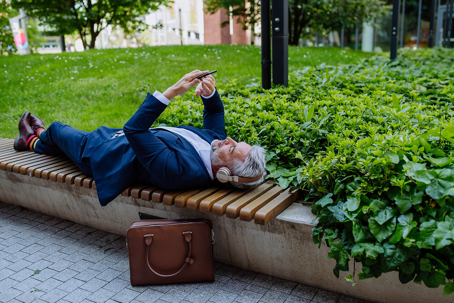 A mature businessman enjoying music in headphones relaxing on bench in city park during break at work.