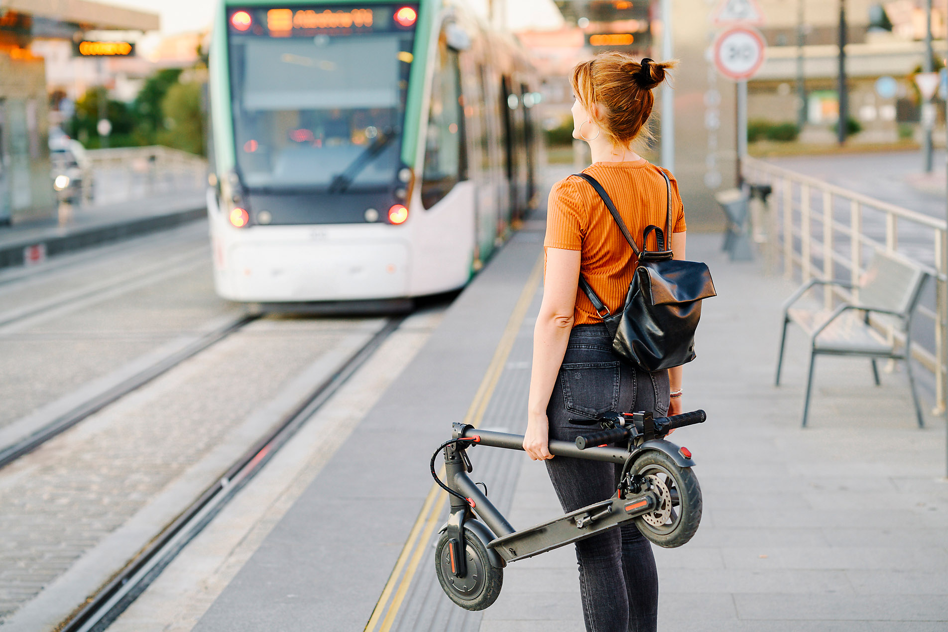 Stock photo, royalty-free, urban, e-mobility, mass transit, back view of woman with backpack and folded electric scooter waiting at streetcar stop, sustainability, smart cities