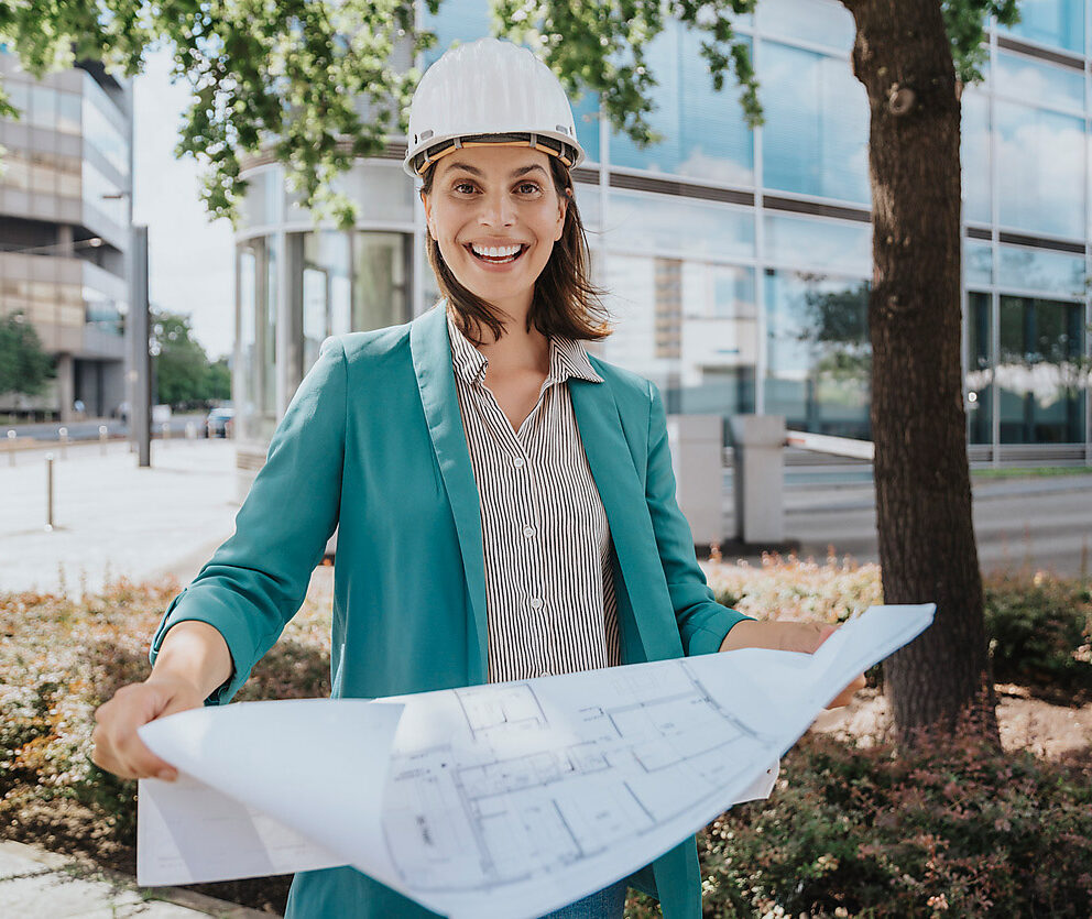 Glückliche Architektin mit Schutzhelm, stehend mit Bauplan, smart cities, westend61, stadtplanung, nachhaltigkeit