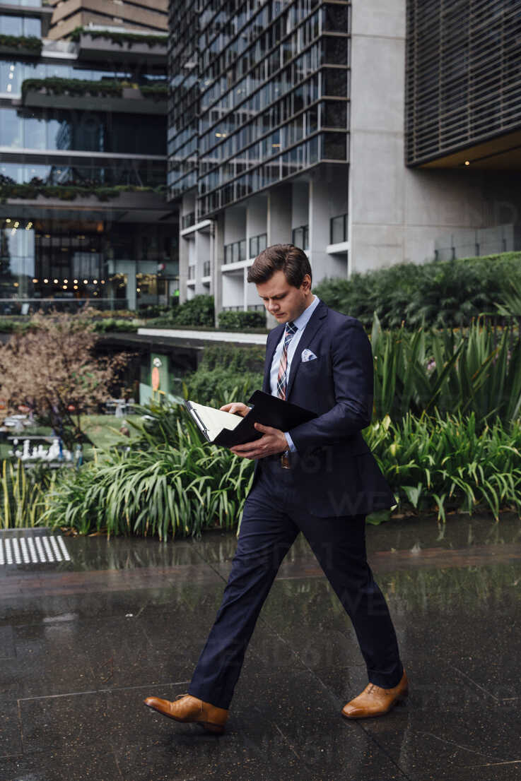 Businessman walking while reading documents on wet footpath in city ...