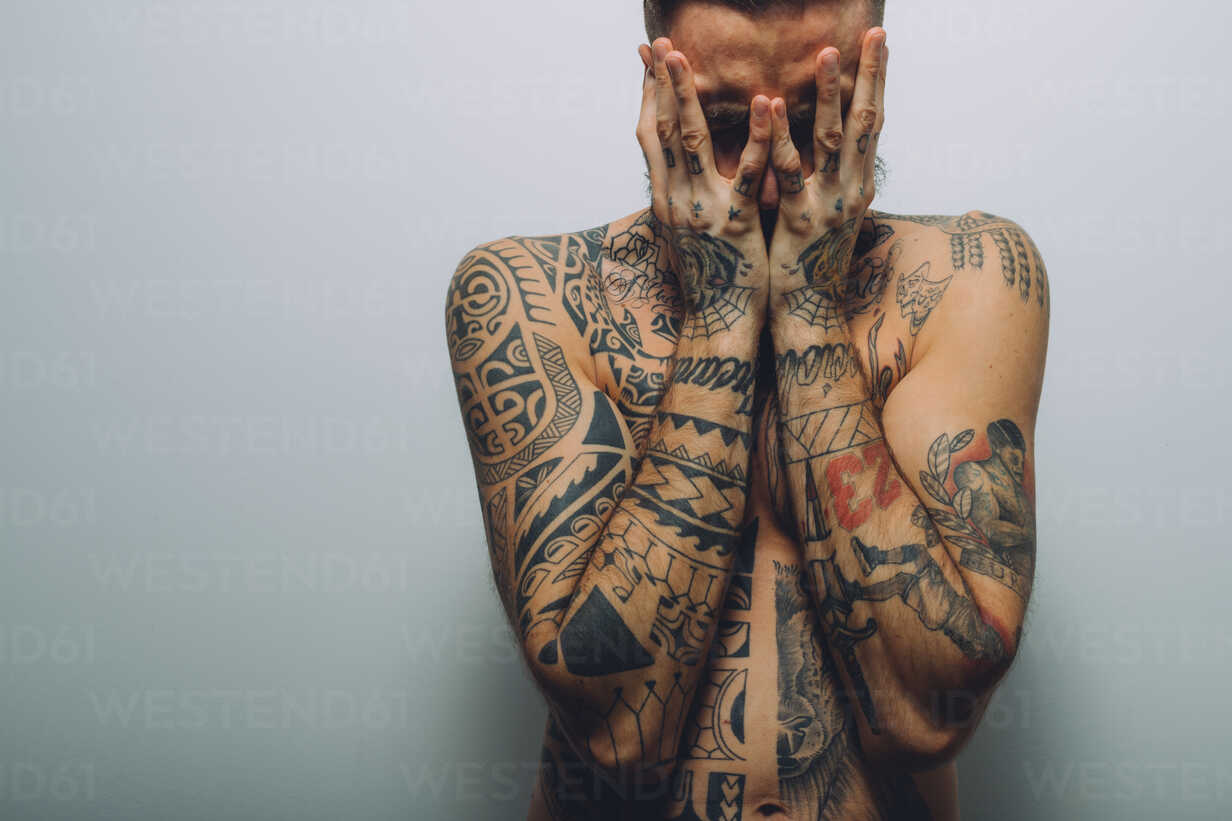 Portrait of young man with beard, covered in tattoos, hands covering face  stock photo
