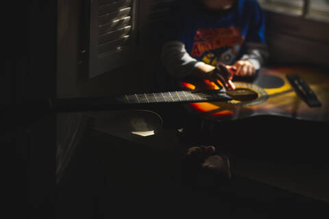 Low section of boy playing guitar while sitting in darkroom at home ...
