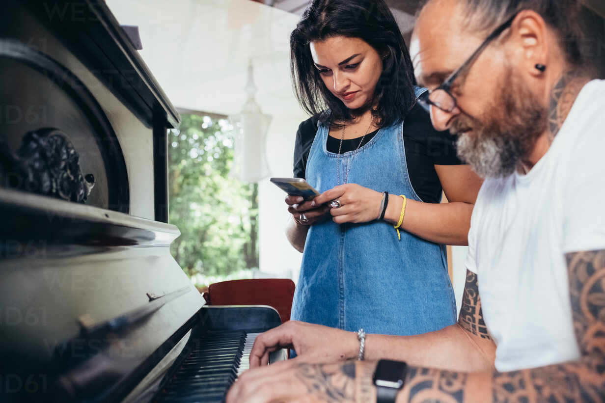 Bearded tattooed man with long hair and glasses playing piano, woman  holding mobile phone listening. stock