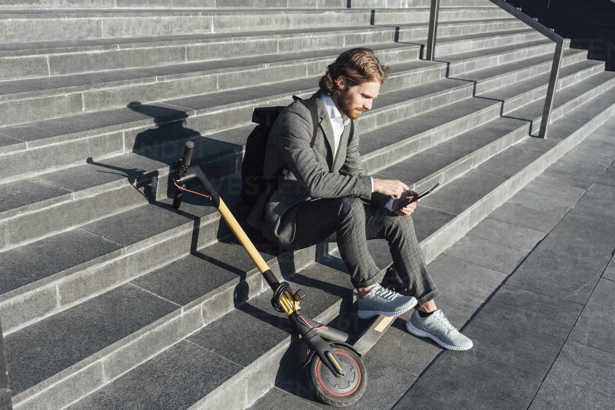 Young businessman with digital tablet and electric push scooter sitting on  steps at downtown during sunny