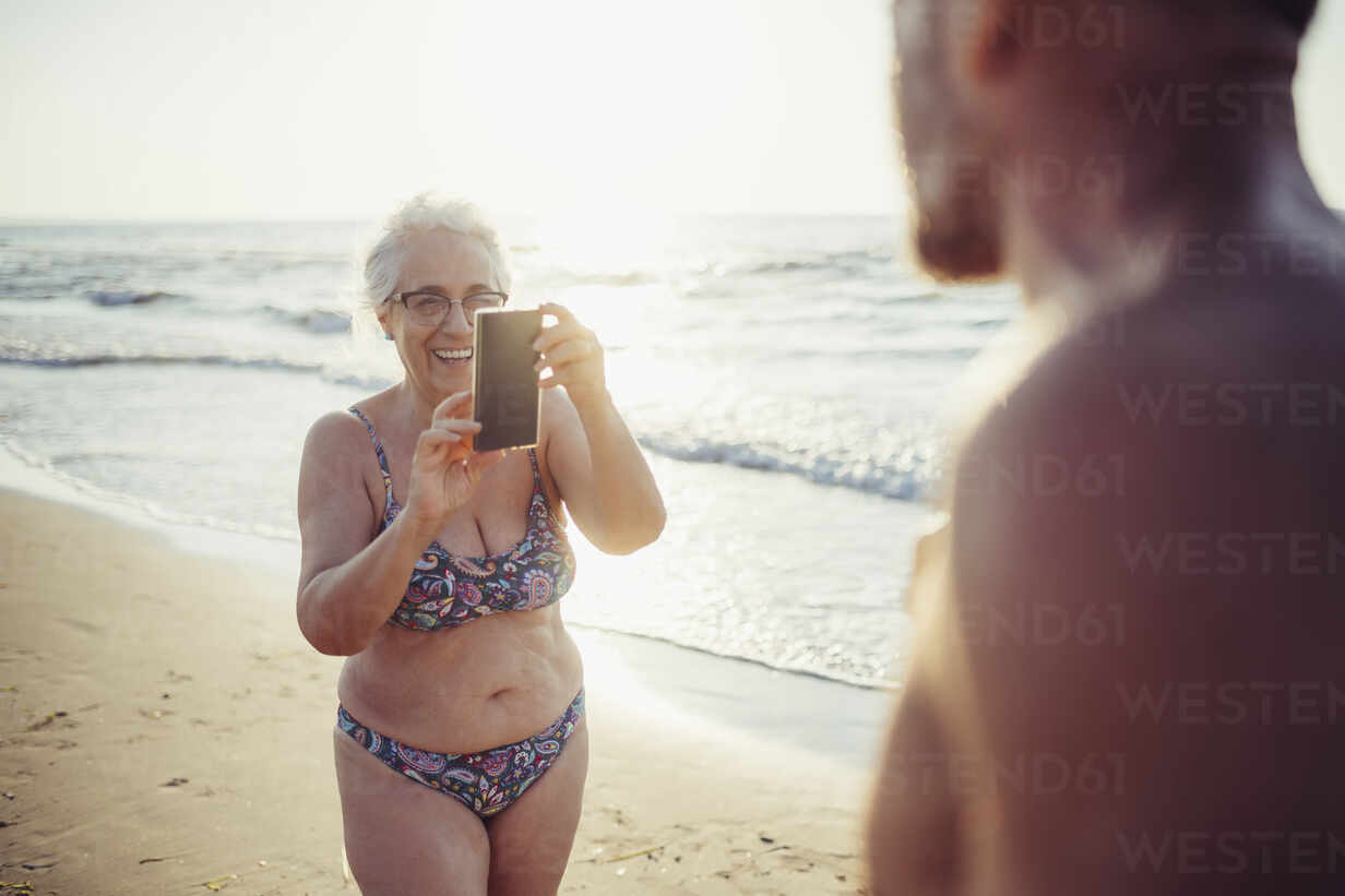senior-woman-in-bikini-taking-photo-of-man-while-standing-at-beach-MEUF02076.jpg
