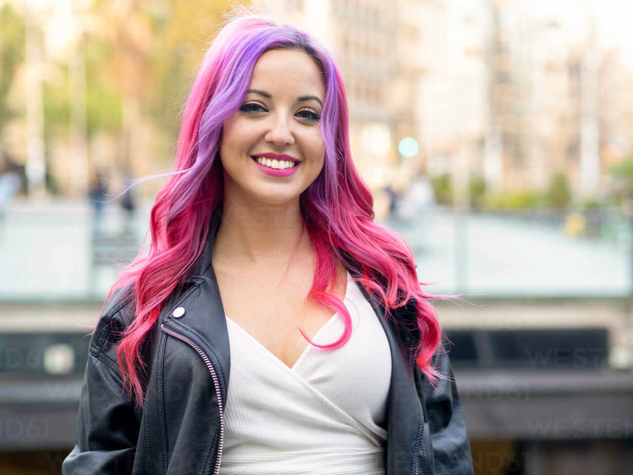 Optimistic confident millennial female with dyed pink hair wearing leather  jacket looking at camera and smiling