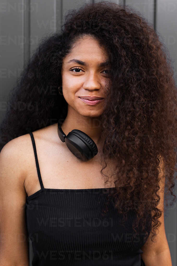 Lovely African American female with long curly hair and headphones on neck  standing in city and