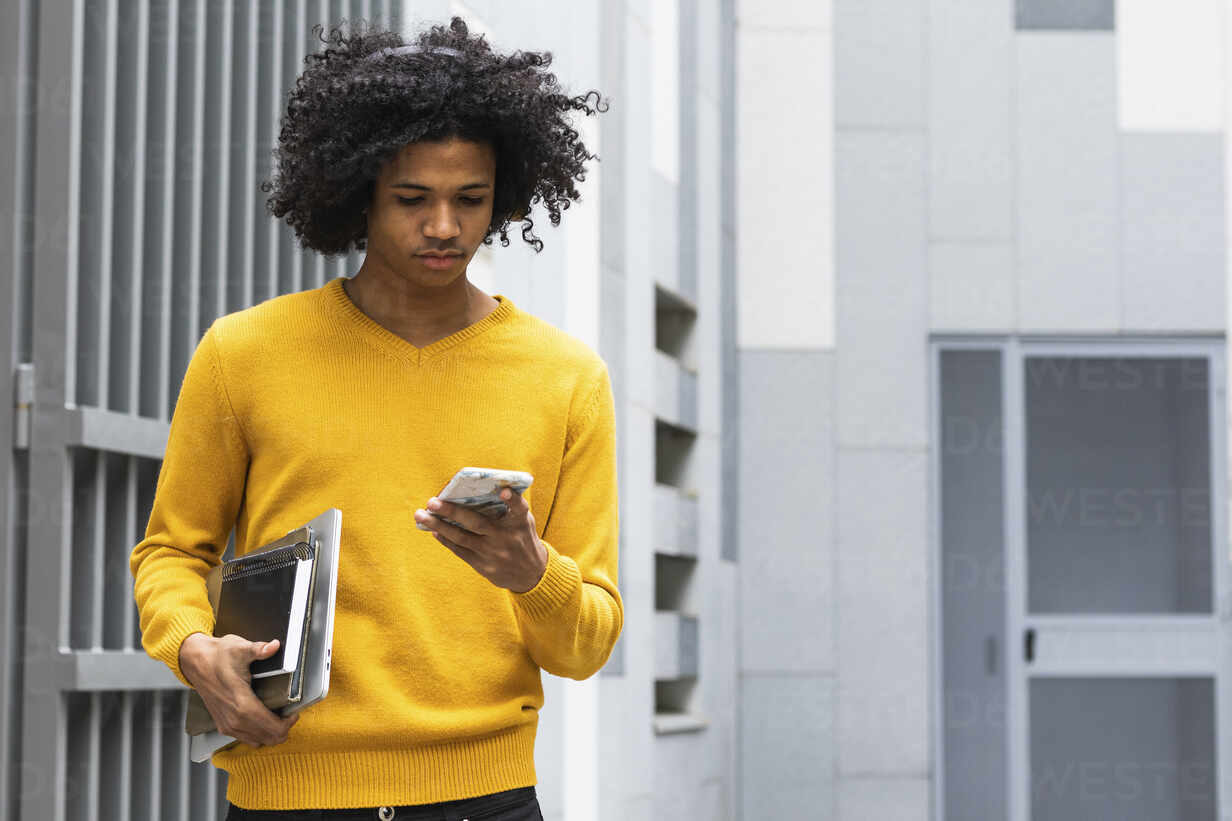 Curly hair boy with books using mobile phone while standing at university  stock photo