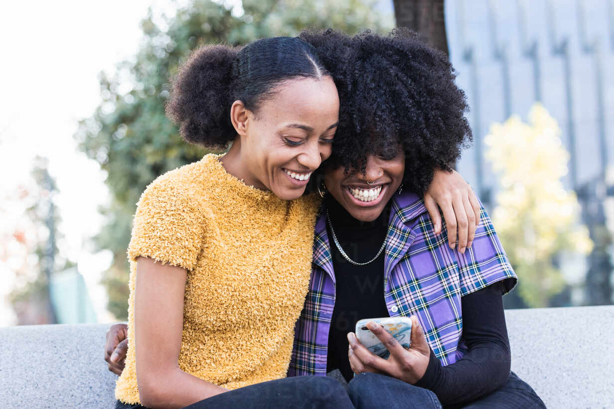 Happy young African American female friends hugging and laughing while  sitting on bench and watching funny