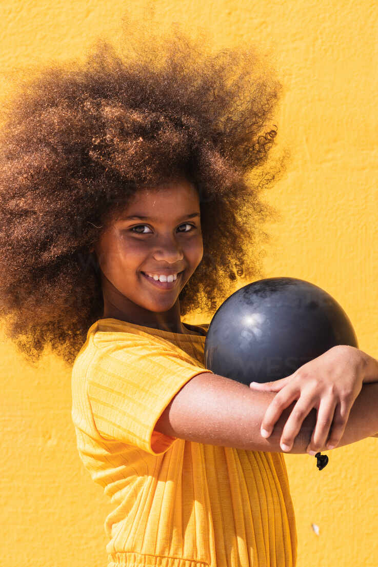 Side view of African American teen girl with curly hairstyle holding black  balloon while standing against