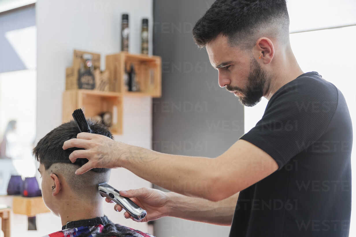 Male barber using machine to cut hair of teenage boy at salon stock photo